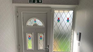 A grey door with a fire exit sign above it, a small window with stained glass, and a doormat on the floor.