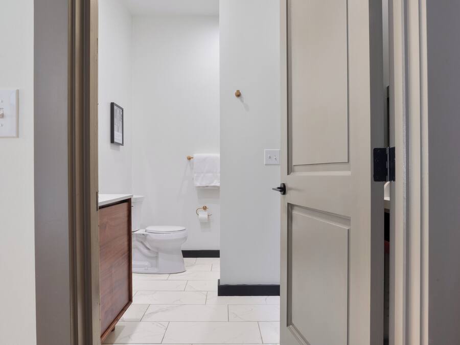 Un couloir avec un sol carrelé de carreaux blancs, une porte fermée sur la droite et des toilettes visibles au fond.