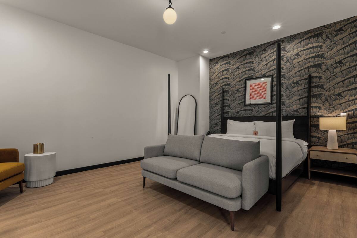 Un dormitorio moderno con un sofá gris, una cama con estructura negra, una mesita de noche con lámpara y una pared decorada con papel tapiz de motivos vegetales.