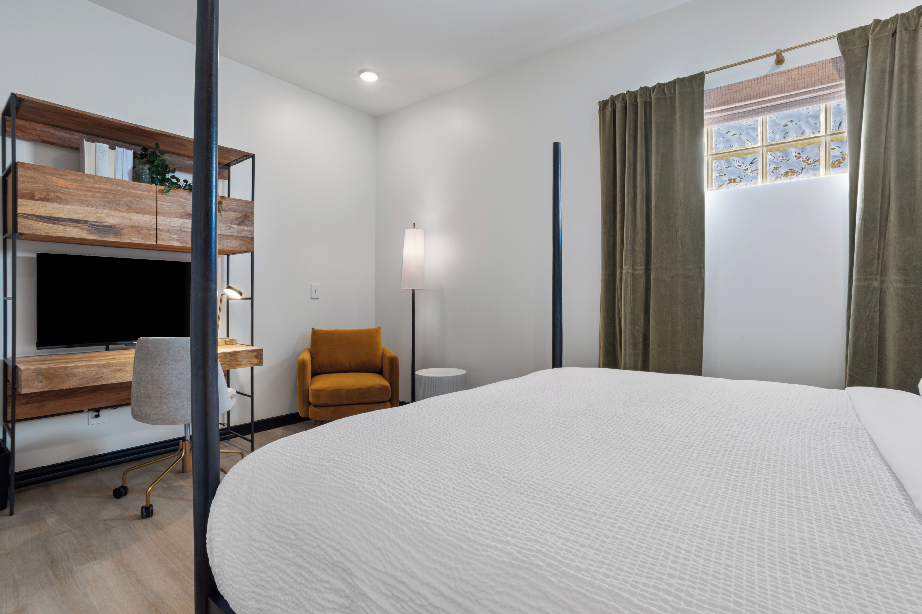 A modern hotel room with a large bed, a wooden desk with a flat-screen TV, a comfortable armchair, and a window with curtains.