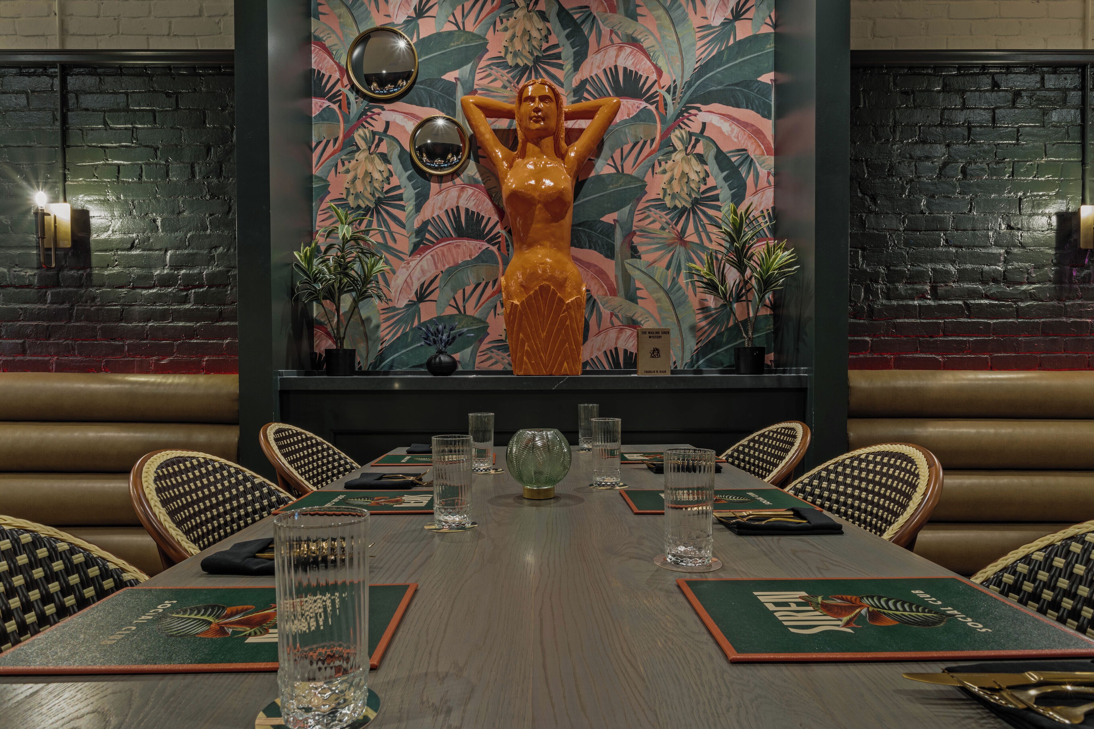 A restaurant with a long wooden table set for dining, a large tropical mural on the wall, and leather booth seating.