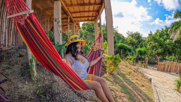 Une personne se détend dans un hamac, sous une structure en bois, avec vue sur un jardin et un chemin de terre.