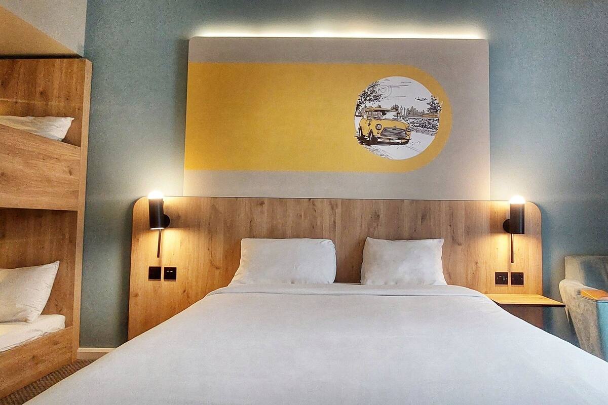 Une chambre d’hôtel avec un lit superposé en bois, un grand lit avec du linge de lit blanc, une œuvre d’art accrochée au mur et un coin salon avec un fauteuil.