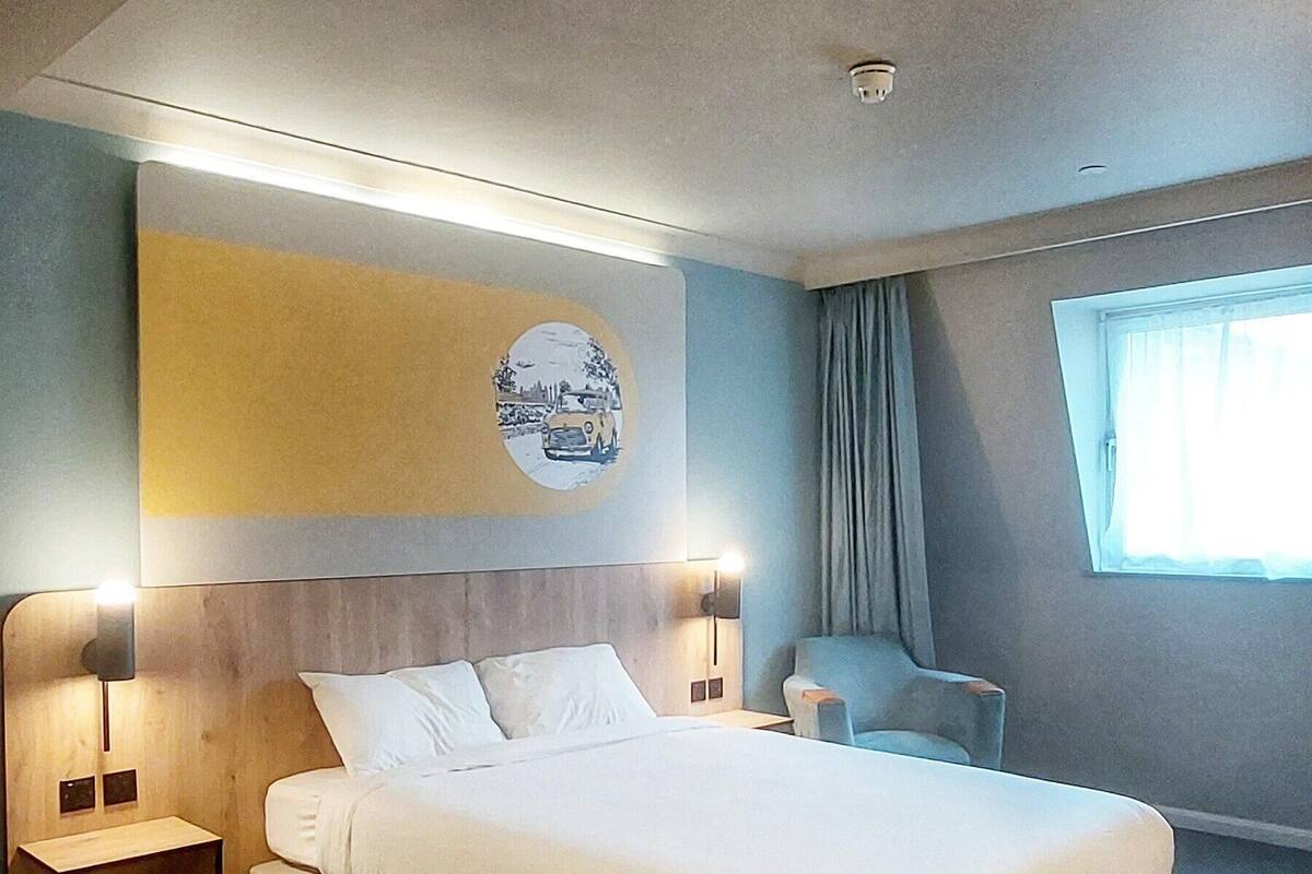 Une chambre d’hôtel moderne avec un grand lit, des tables de chevet, une œuvre d’art accrochée au mur, une fenêtre avec des rideaux et un luminaire au plafond.