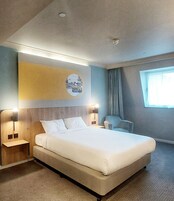 A modern hotel room with a large bed, bedside tables, a wall-mounted artwork, a window with curtains, and a ceiling light.