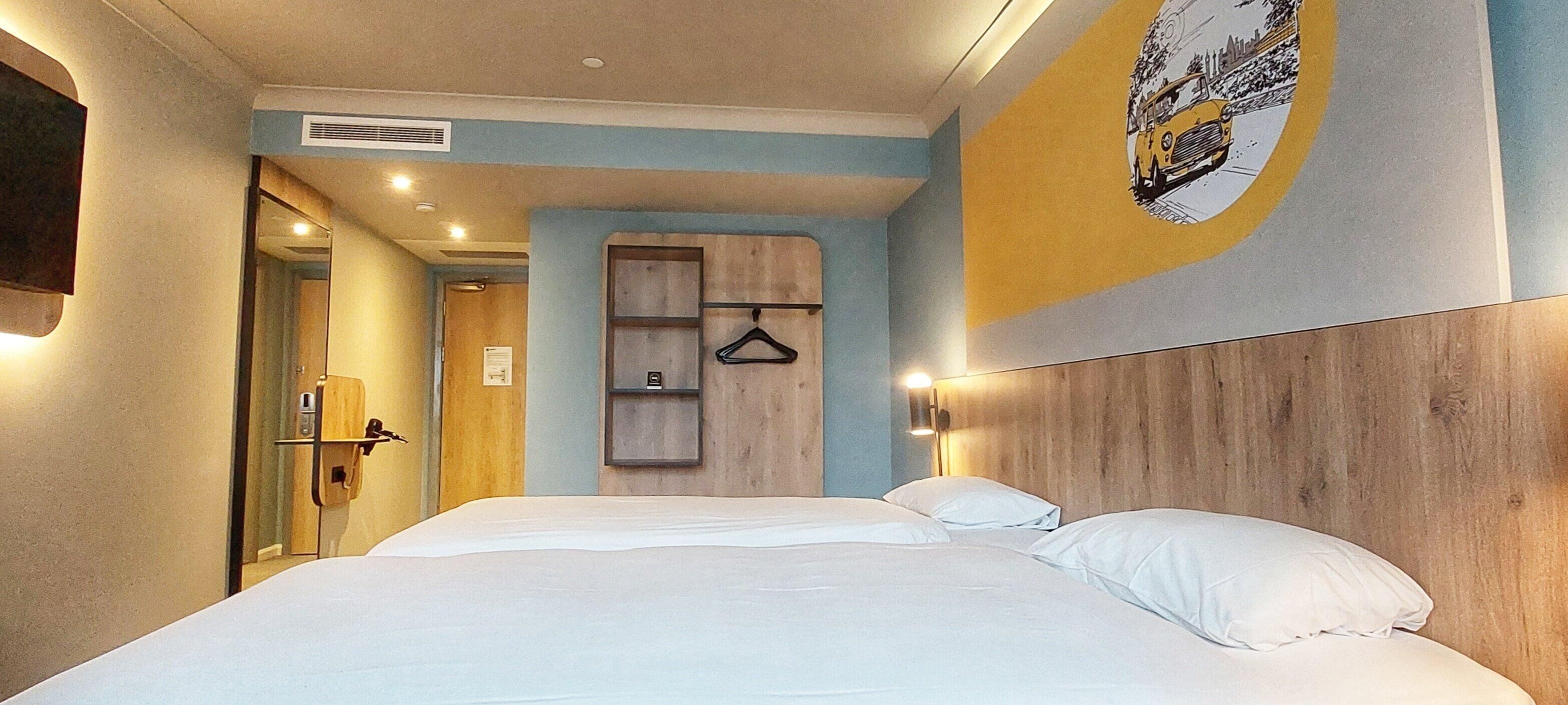 A hotel room with a large bed, a wooden headboard, a wall-mounted coat rack, and a door leading to a bathroom.