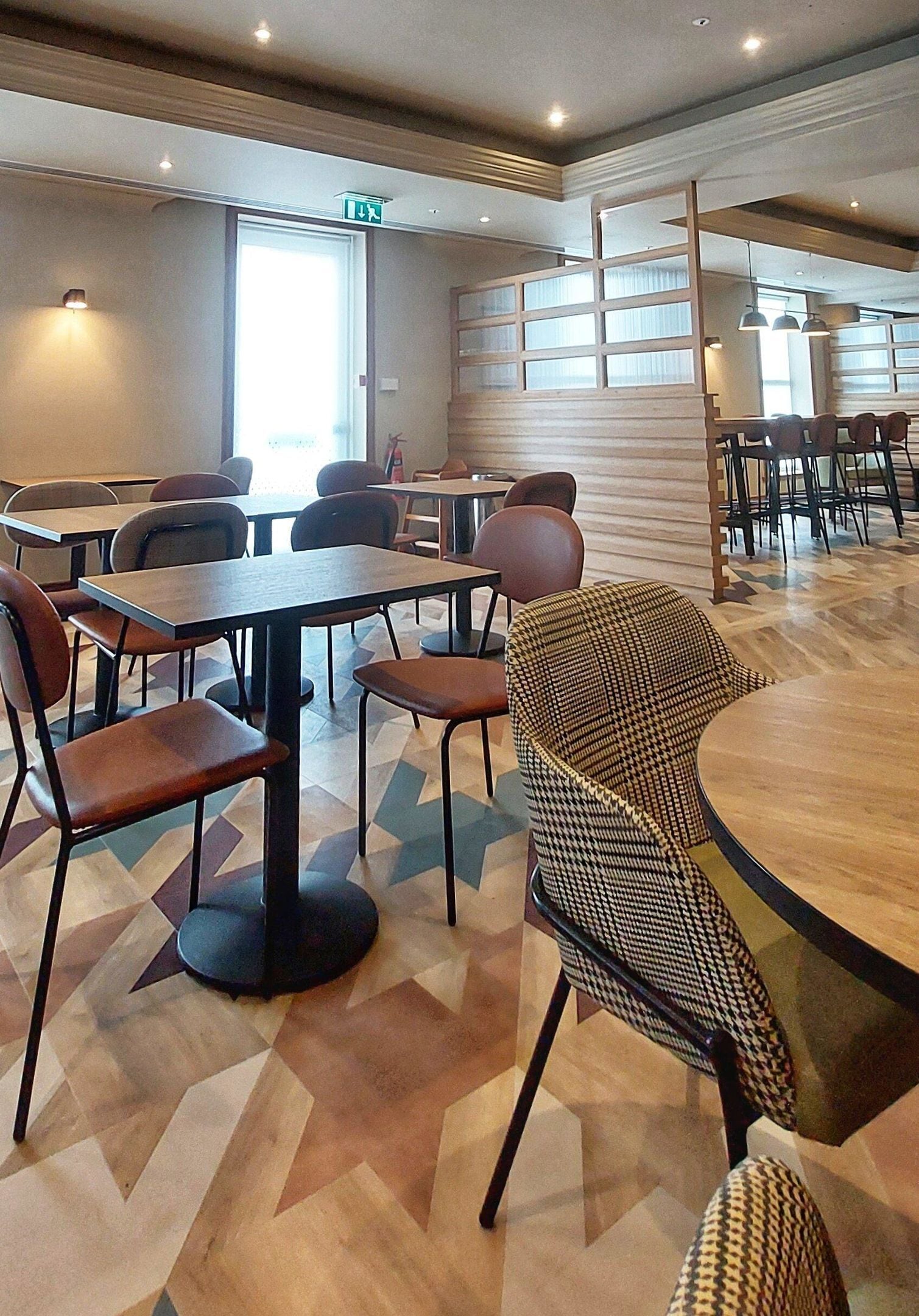 A modern dining area with multiple tables and chairs, a patterned floor, and a large window allowing natural light.