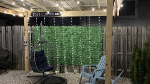 A covered outdoor seating area with a wooden pergola, a green wall with lights, and two chairs.