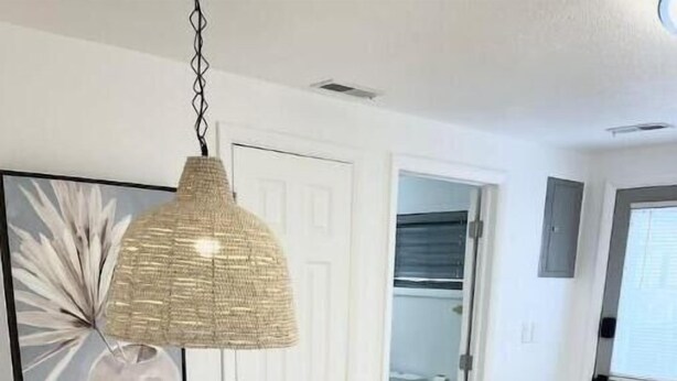 A dining area with a round table and white chairs, a hanging woven light fixture, and a door leading to a bathroom.