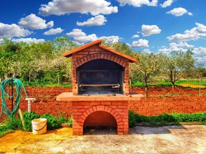 A brick outdoor oven with a sloped roof, surrounded by a garden with trees and a fence.