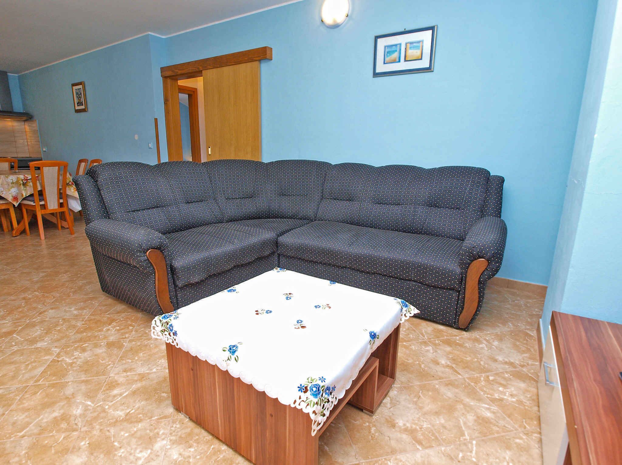 A living room with a sectional sofa, a small table with a floral tablecloth, a dining area with chairs and a table, and a door leading to another room.