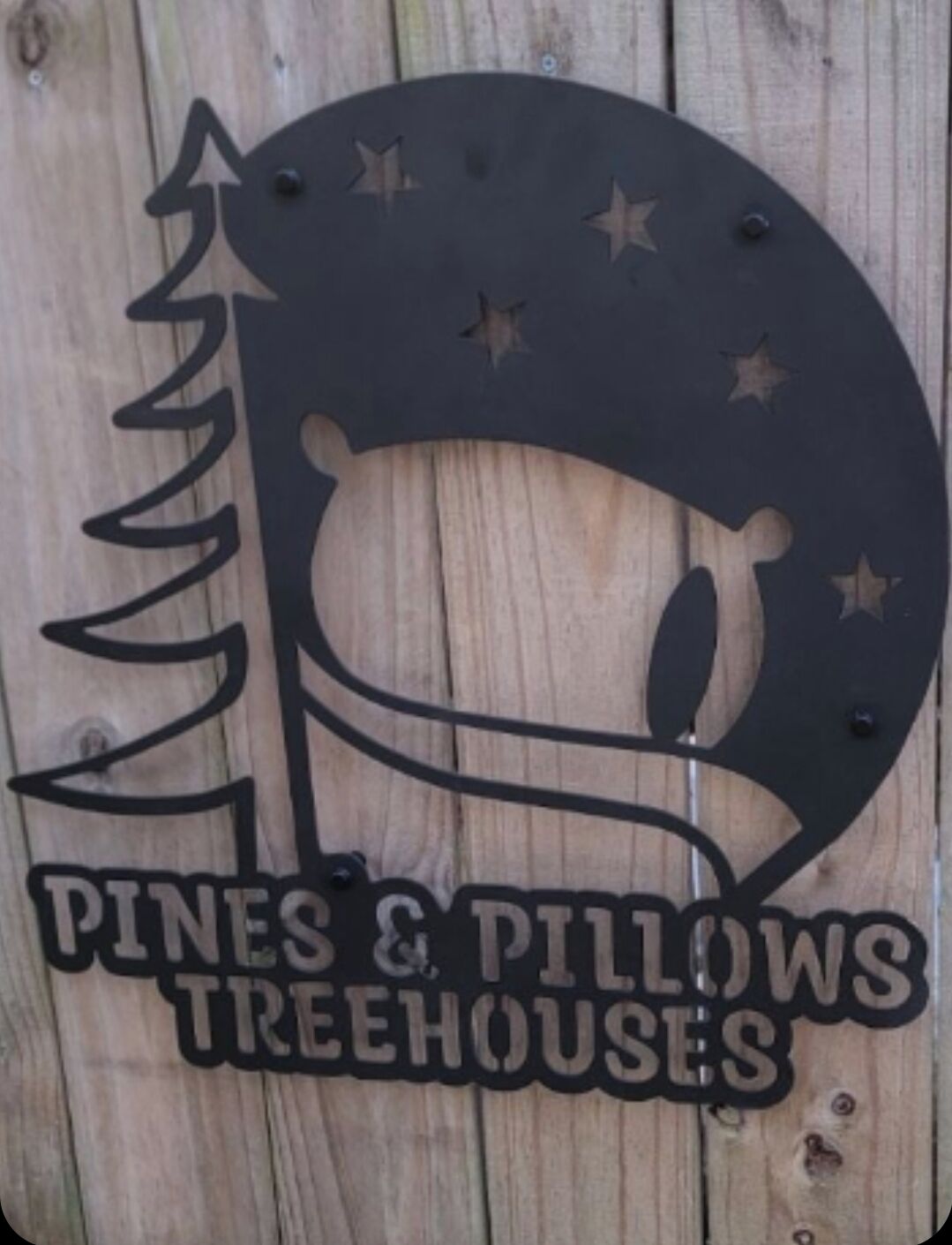 A metal sign with a crescent moon and stars, and the text "Pines & Pillows Treehouses".