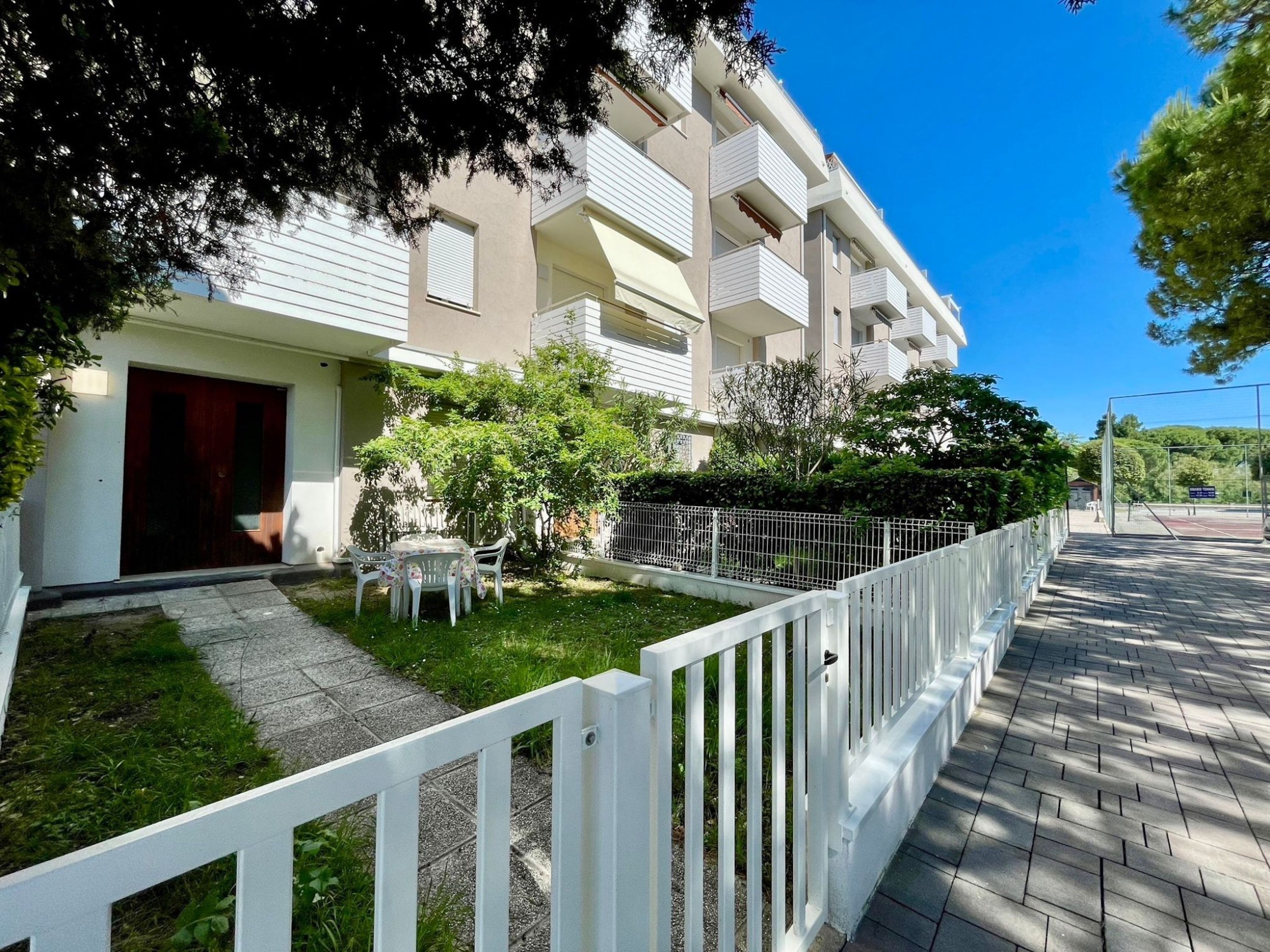 A modern apartment building with a white exterior, a small garden with a table and chairs, and a white fence along the sidewalk.