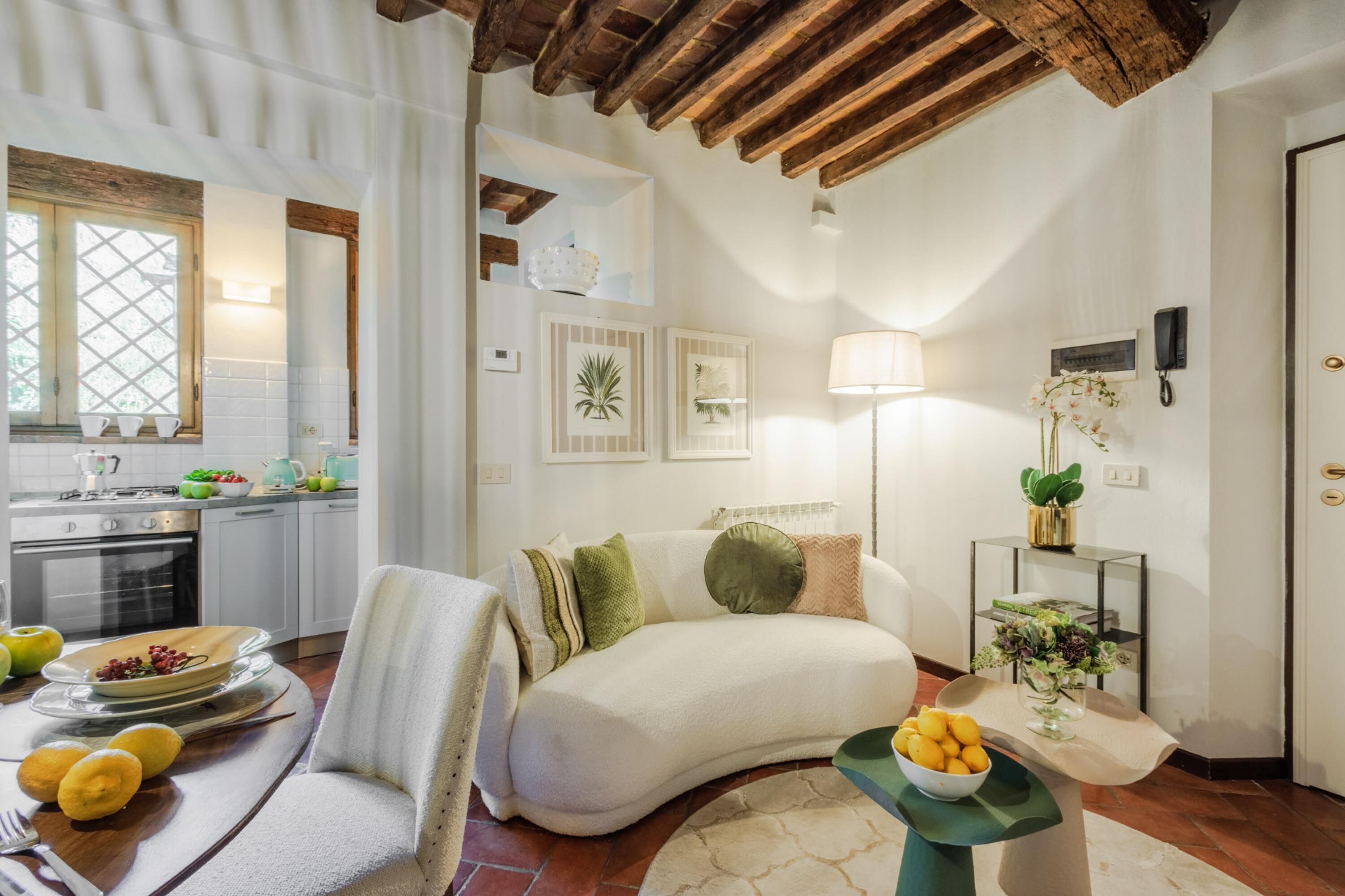 Un salon chaleureux avec un canapé, une table basse et un coin repas avec une table dressée pour deux personnes. Au fond, on aperçoit une cuisine avec une fenêtre et une plante posée sur le comptoir.