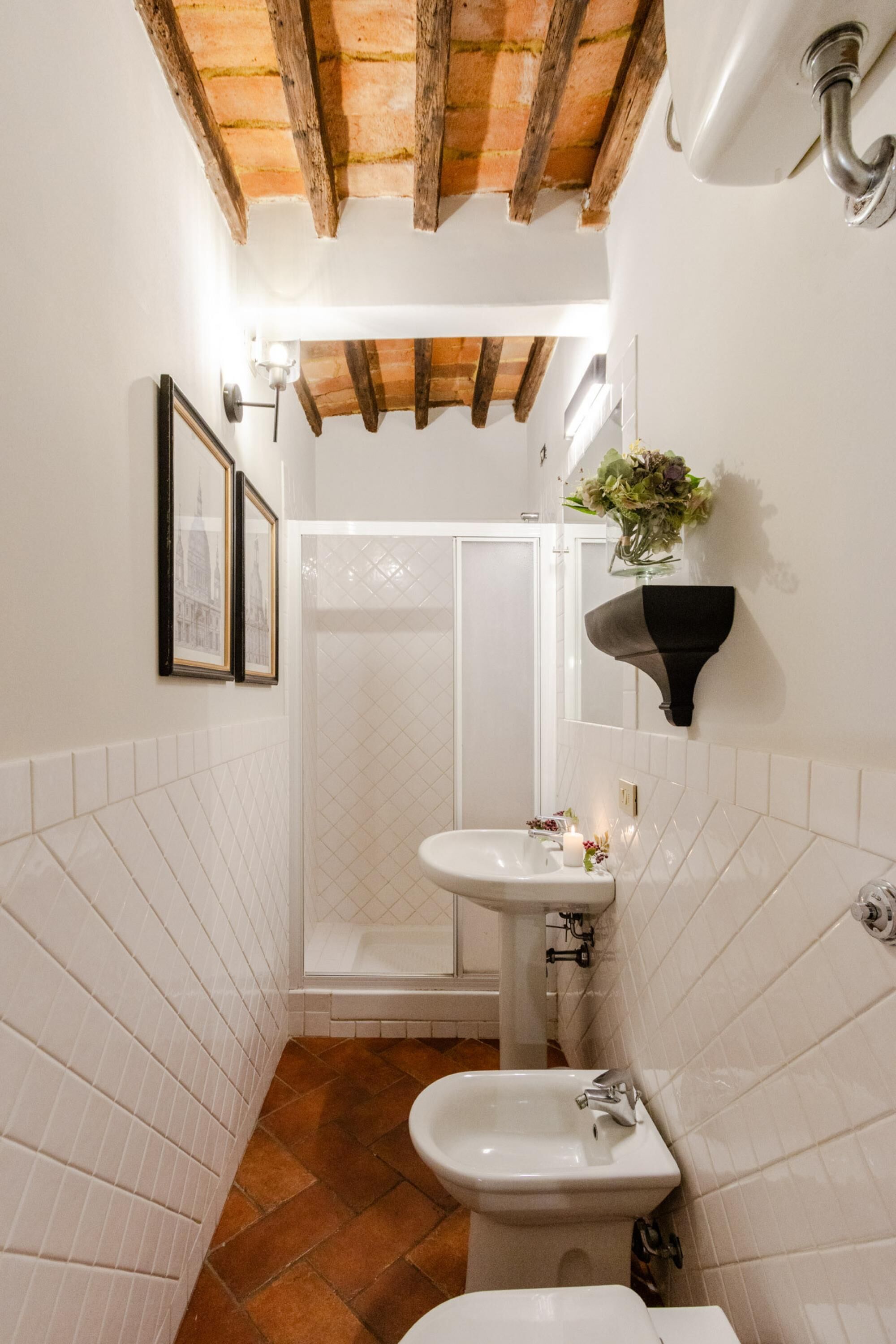 Un bagno stretto con pareti rivestite di piastrelle bianche, soffitto in legno, lavandino bianco, bidet e doccia.