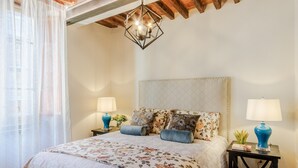Un dormitorio con una cama grande, ropa de cama floral, dos lámparas de noche, una lámpara de techo geométrica y una ventana con cortinas de gasa.