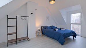 Een slaapkamer met een schuine kap, een bed met blauw beddengoed, een nachtkastje met een lamp en een hangrek.