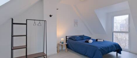Une chambre avec un plafond en pente, un lit avec une literie bleue, une table de chevet avec une lampe et un porte-vêtements suspendu.