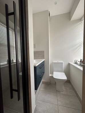 Bathroom features a toilet, sink, and window with blinds, set against a backdrop of light-colored tiles.