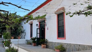 Un edificio blanco con detalles en rojo y techo de tejas, que cuenta con un pequeño porche con plantas en macetas y una puerta.