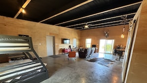 A spacious room with a high ceiling, a large window, a mounted television, and a ceiling fan. - Sprague Ranch Cabin: Hill Country Views, Mesquite Firepit, Trails & Pond (Lometa)