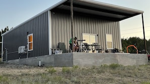 A single-story metal building with a covered porch, a concrete foundation, and a small outdoor seating area with tables and chairs. - Sprague Ranch Cabin: Hill Country Views, Mesquite Firepit, Trails & Pond (Lometa)