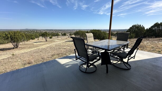 An outdoor patio with a table and chairs, overlooking a dry landscape with sparse trees and a distant view of a city. - Sprague Ranch Cabin: Hill Country Views, Mesquite Firepit, Trails & Pond (Lometa)