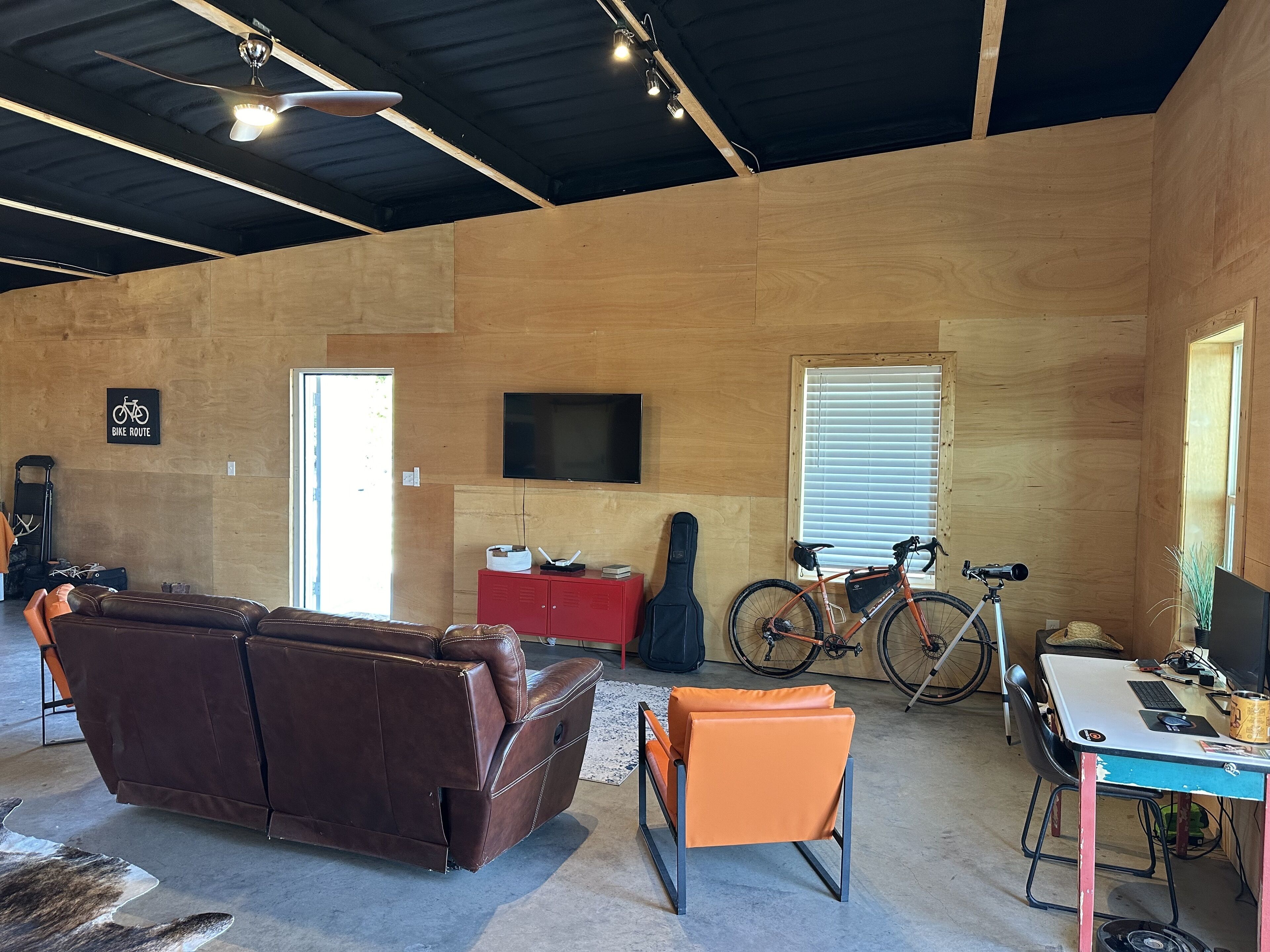 Une pièce spacieuse avec un canapé en cuir marron, un fauteuil orange, un meuble rouge, un vélo et un téléviseur fixé au mur.