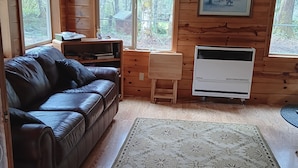 A cozy cabin interior with a leather sofa, a small wooden table, a white cabinet, and a large window offering a forest view. - Lower Twin Cabin - 34 beautiful private acres of forest, meadows and riverfront. (Twin)