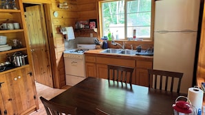 A wooden cabin kitchen with a stove, sink, and refrigerator. There is a wooden table with chairs, a window with a view of trees, and various kitchen items on the shelves. - Lower Twin Cabin - 34 beautiful private acres of forest, meadows and riverfront. (Twin)