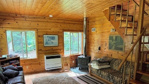 A cozy cabin interior with a wooden interior, a fireplace, a sofa, a bookshelf, and a staircase. - Lower Twin Cabin - 34 beautiful private acres of forest, meadows and riverfront. (Twin)