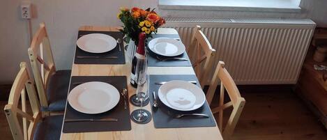A dining area with a wooden table set for four, a vase with flowers, and a view of a landscape through a window.