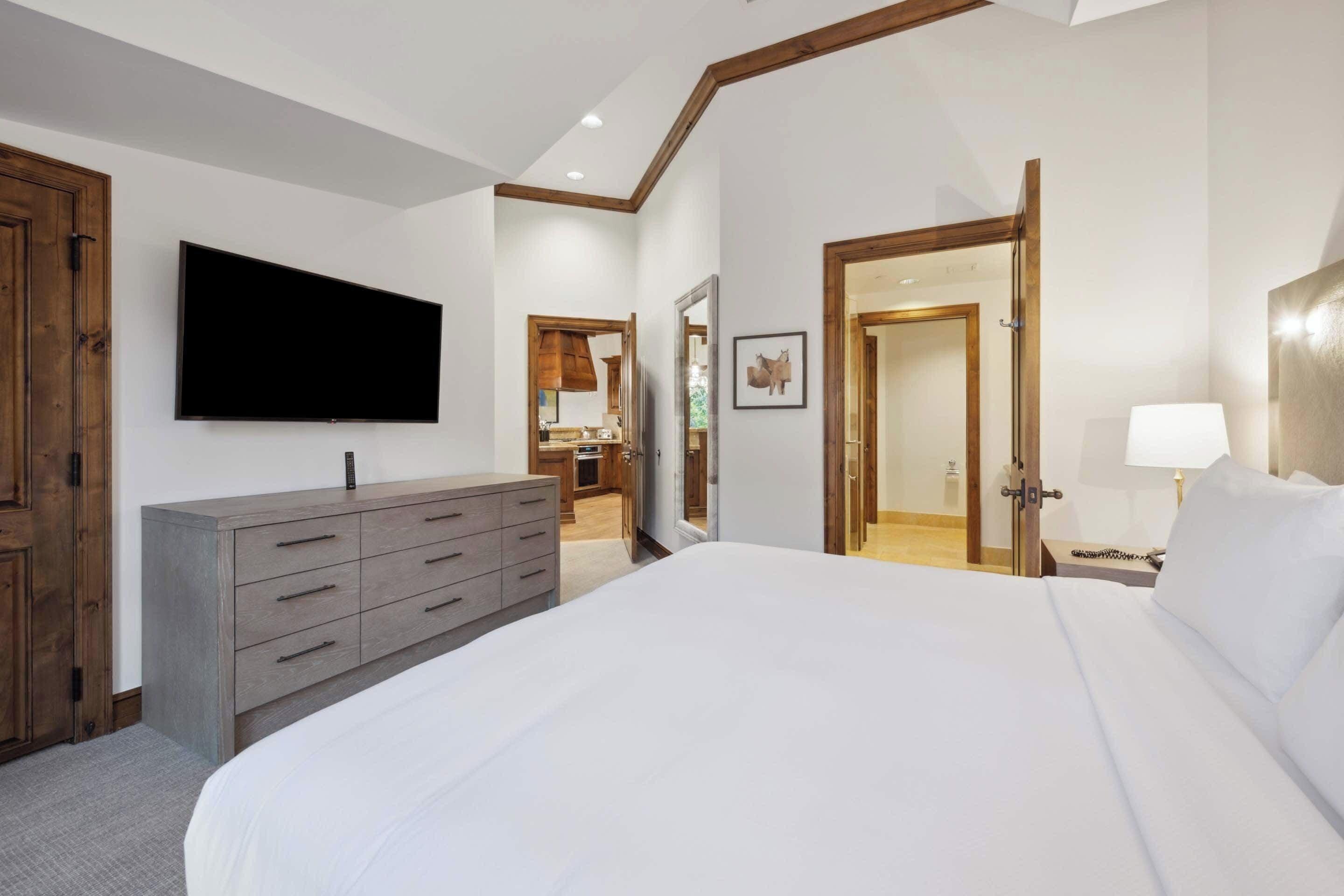 Een slaapkamer met een groot bed, een aan de muur bevestigde televisie, een houten dressoir en een deur naar een badkamer.