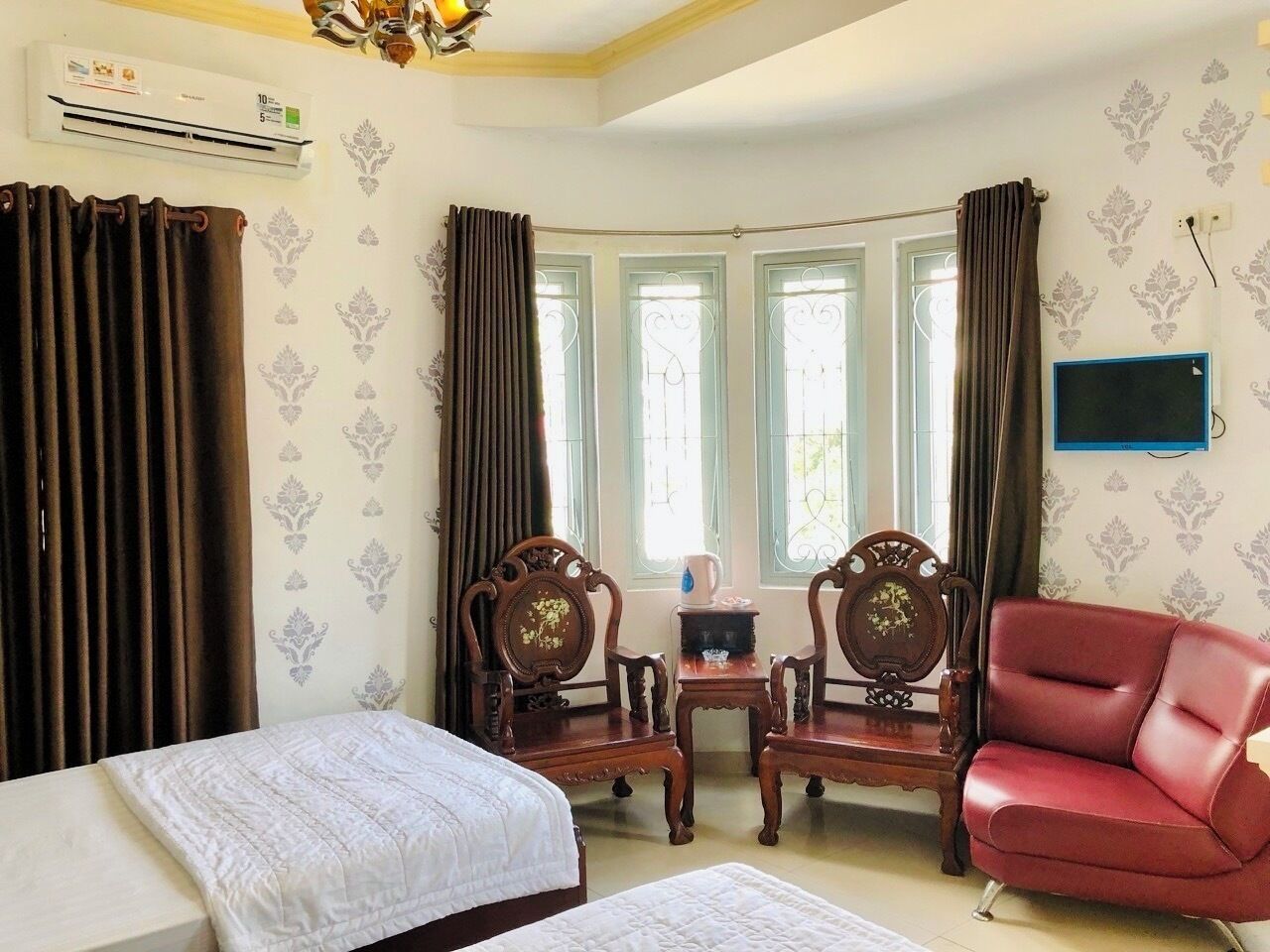 Une chambre d’hôtel avec deux lits, un fauteuil rouge, des chaises en bois, une petite table et un téléviseur fixé au mur.
