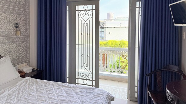 A hotel room with a bed, a desk, a chair, a TV, and a balcony with a view of greenery. - Hong Tam Hotel (Ho Chi Minh City)