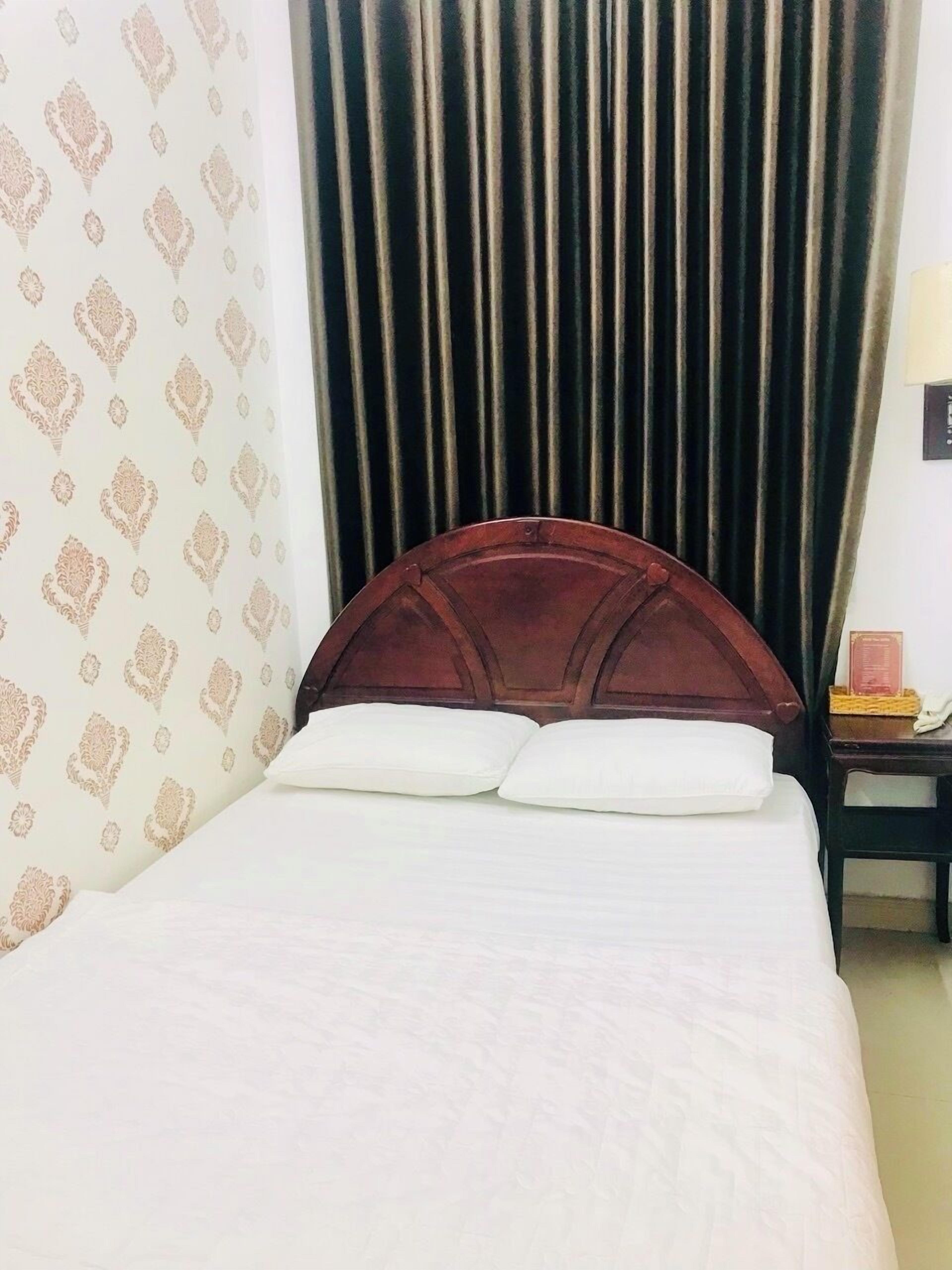 Ein ordentlich bezogenes Bett mit Holz-Kopfteil, einer gemusterten Wand und einem dunklen Vorhang.
