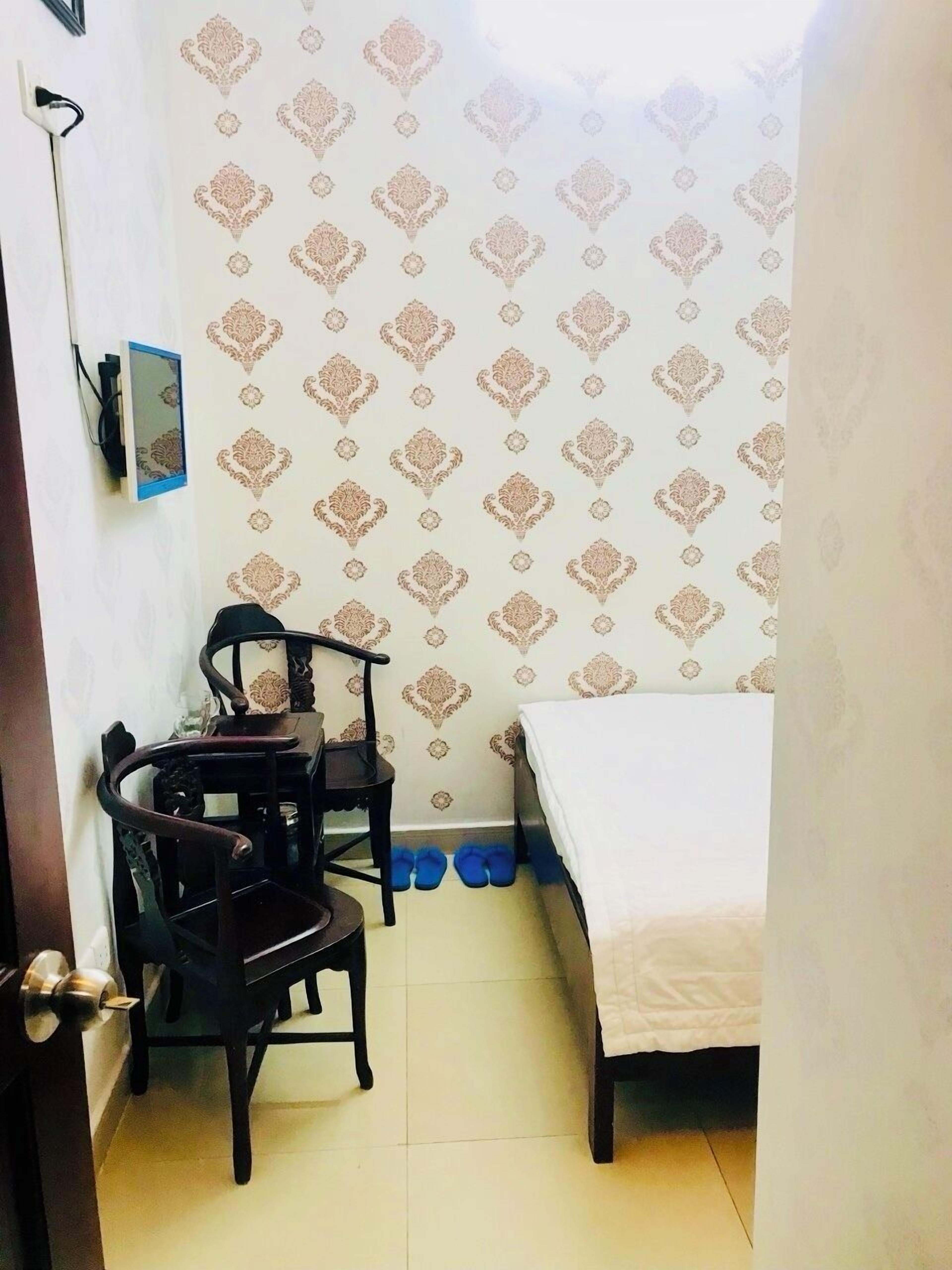 Una habitación pequeña y estrecha con una cama, un escritorio y una silla. Las paredes tienen un papel tapiz estampado, y hay un estante con objetos encima.