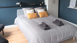 Un dormitorio con cama, ropa de cama con estampado gris, dos almohadas, una mesita de noche, una lámpara y un armario.