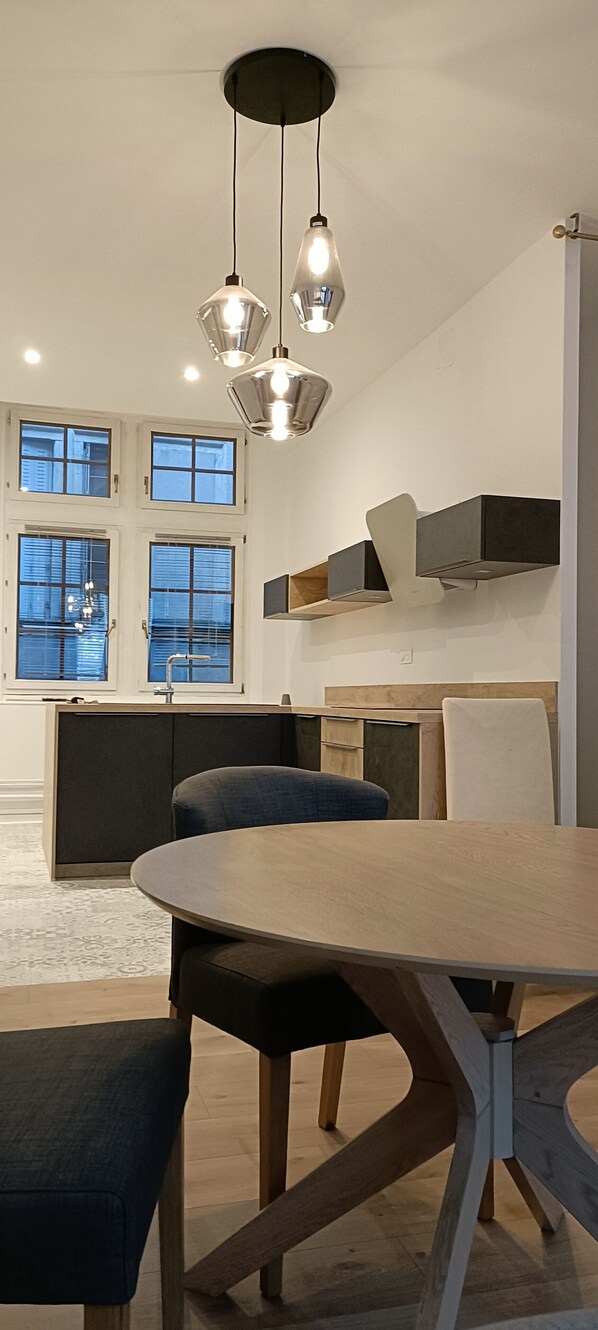 A modern kitchen with a dining area, featuring a wooden table and chairs, pendant lighting, and a view of the city through large windows.