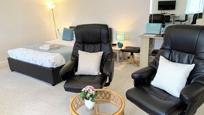 Moderni hotellihuone, jossa on sänky, kaksi mustaa nahkaista nojatuolia, pieni pyöreä pöytä maljakko täynnä kukkia, työpöytä tietokoneella ja seinälle asennettu televisio.