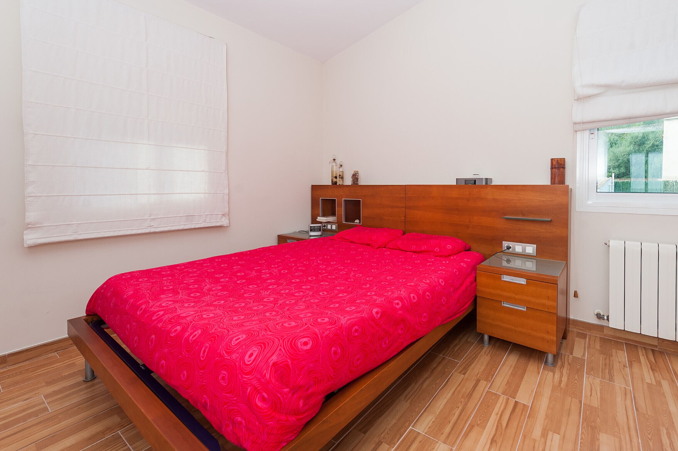 Una camera da letto con un letto di legno, un comodino, un cassetto, una finestra con persiane e un termosifone.