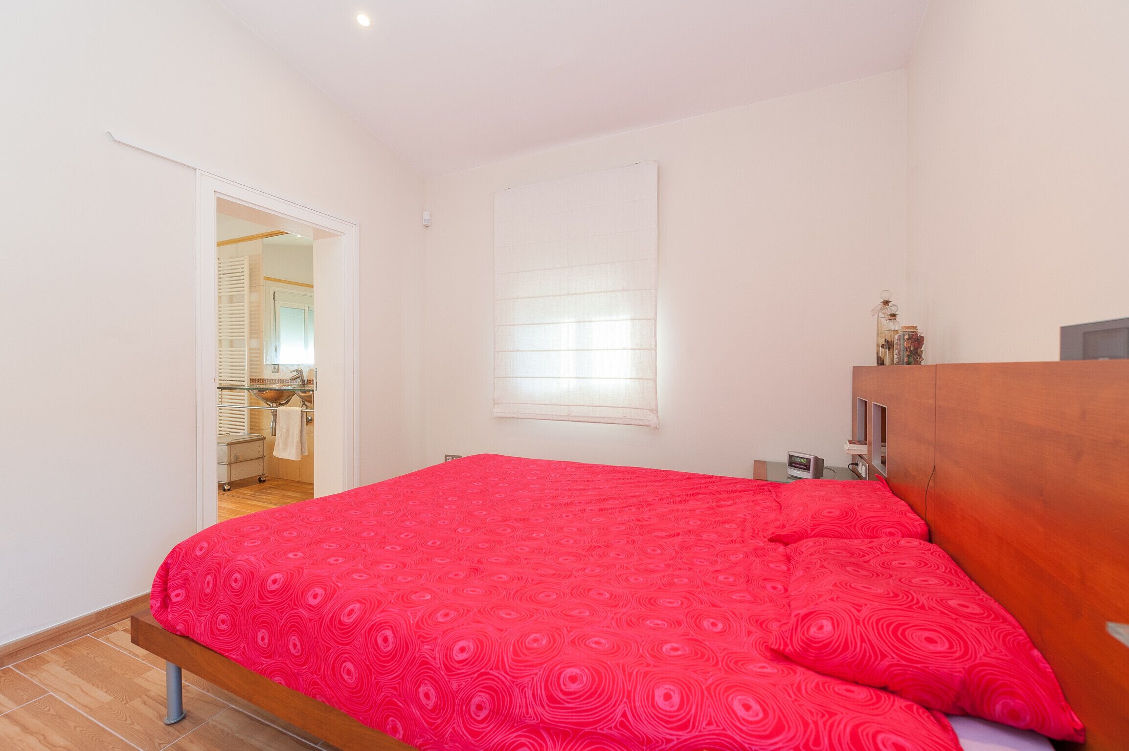 A bedroom with a red patterned bedspread, a wooden headboard, a small bedside table with a lamp, and a window with blinds.