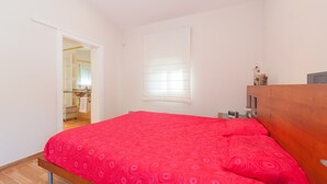 A bedroom with a red patterned bedspread, a wooden headboard, a small bedside table with a lamp, and a window with blinds. - Sunset Villa Romani (Ca´n Picafort)