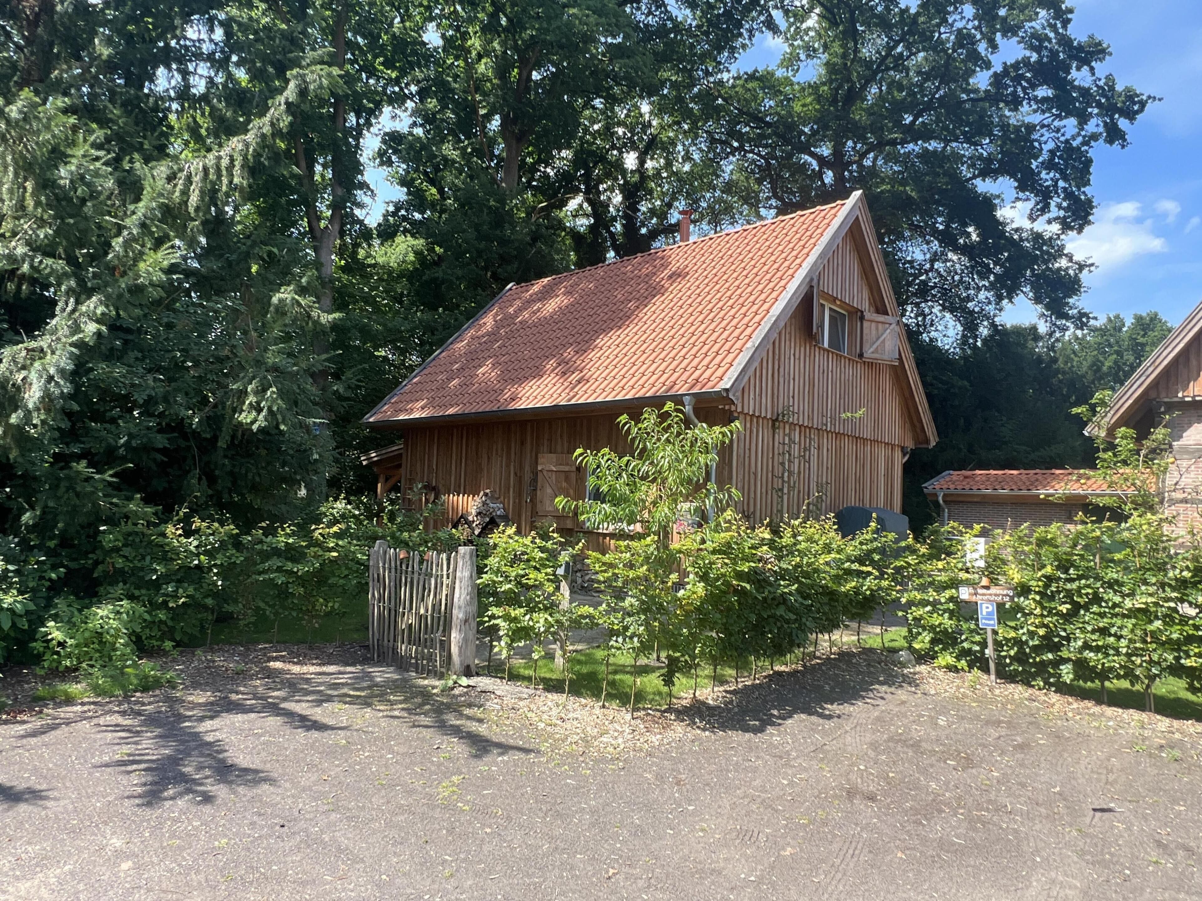 Una baita di legno con tetto in tegole rosse, immersa tra alberi fitti e un vialetto sterrato.