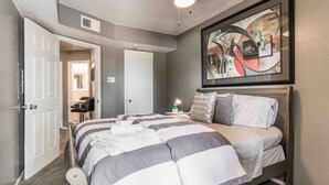 A bedroom with a bed, a framed abstract painting, a bedside table with a lamp, and a door leading to another room. - Cute One Bedroom Apartment w. Balcony! (Dallas)