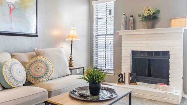 A living room with a beige sofa, patterned cushions, a side table with a lamp, a fireplace, and a window with blinds. - Cute One Bedroom Apartment w. Balcony! (Dallas)
