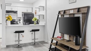 Moderni keittiö, jossa on valkoiset kaapit, musta jääkaappi ja baaritiski, jossa on kaksi jakkaraa. Seinälle on asennettu litteänäyttöinen televisio, ja sen alla on puinen hylly.