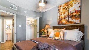 Une chambre avec un grand lit, une table de chevet avec une lampe, un tableau accroché au mur et une salle de bain visible à travers une porte ouverte.