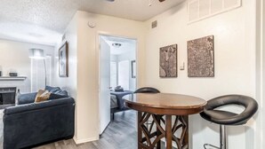 A compact living space with a dark-colored sofa, a wooden dining table, and two black bar stools. A fireplace is visible in the background, and the room has a white ceiling with a vent. - STAY AWHILE IN THIS COZY 1 BED APT! (Dallas)