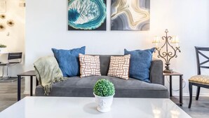 A modern living room with a grey sofa, patterned cushions, a white coffee table with a green plant, and abstract wall art. - N. Dallas Updated 1 Bedroom Apartment (Dallas)